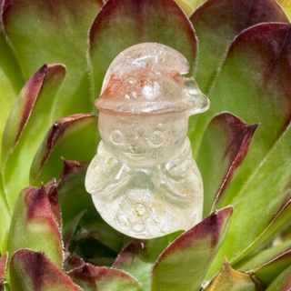 Clear crystal snowman carving nestled in a green succulent with burgundy-tipped leaves.